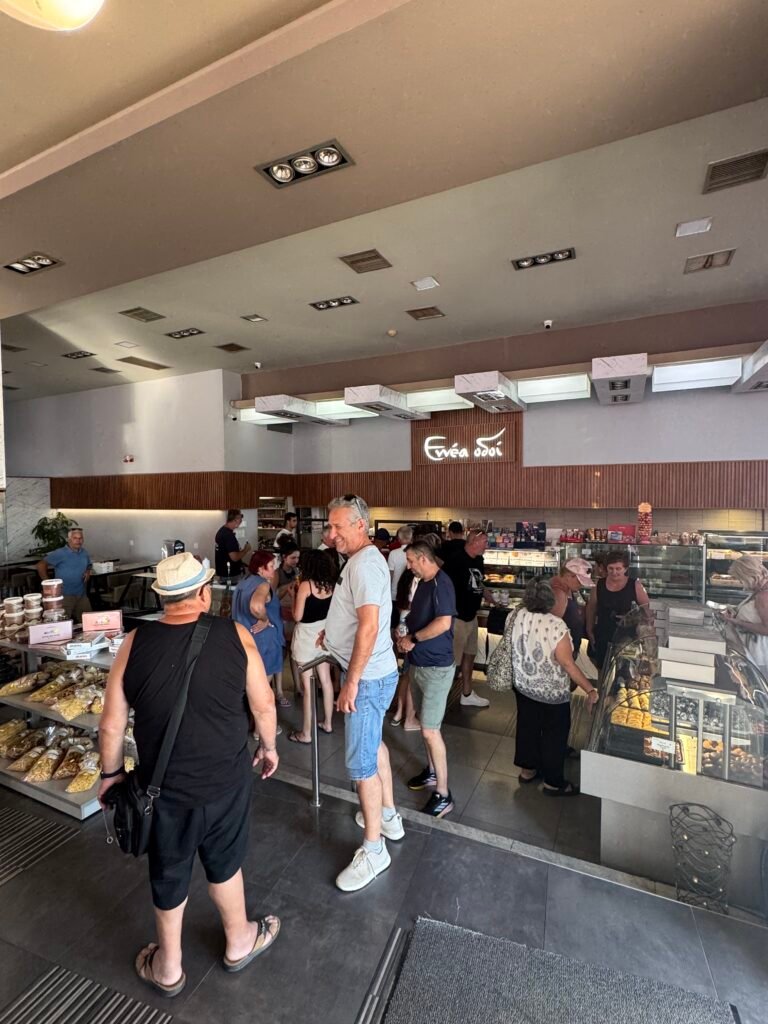 Busy bakery/café interior with a long line of customers and a glass display case filled with pastries on the right.