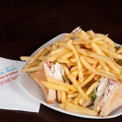 Plate of crispy french fries piled on a white plate with triangular sandwiches (ham, cheese, lettuce) on the side; a napkin lies nearby on a dark table edge.