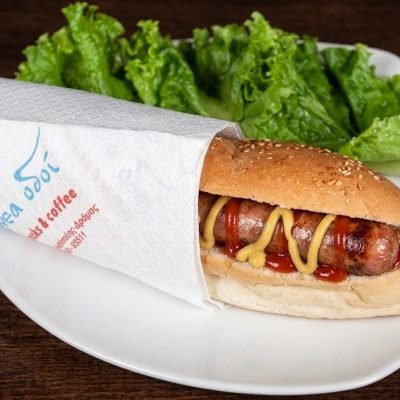 Hot dog in a sesame bun with mustard and ketchup on a white plate, partially wrapped in a napkin with lettuce on the side.